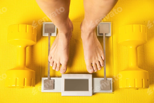 Preview: Female feet on the modern weighing scale with a yellow dumbbells and fitness mat