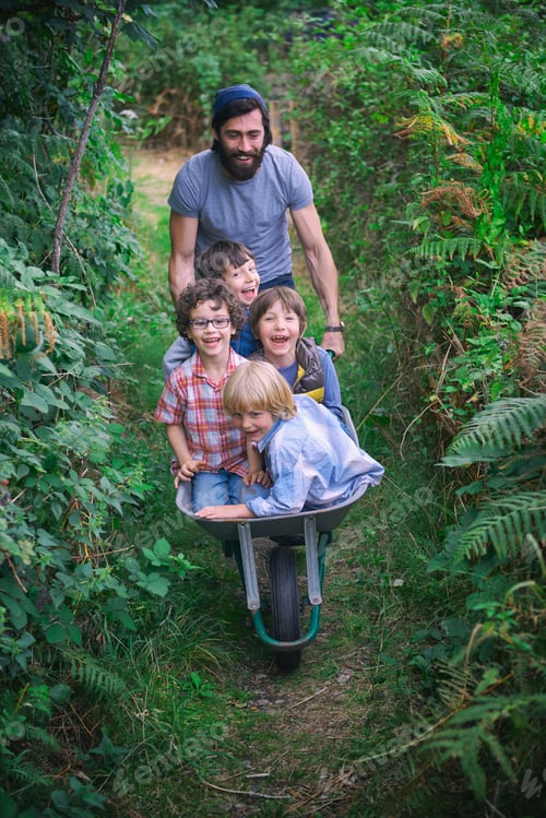 Preview: Mid adult man pushing wheelbarrow with smiling boys in garden