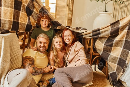 Preview: joyful parents and kids laughing under blanket hut in living room, playing together at home