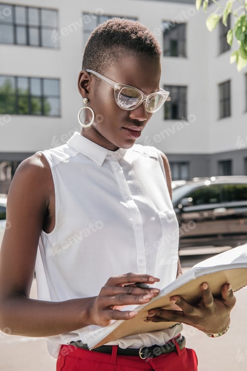 Preview: stylish attractive african american businesswoman looking at documents on street