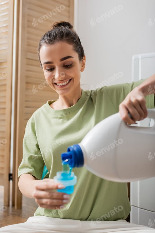 Preview: Cheerful brunette woman pouring blue washing liquid in laundry room