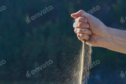 Preview: Transience, sand runs through clenched fingers on dark blurred background outdoors copy space.