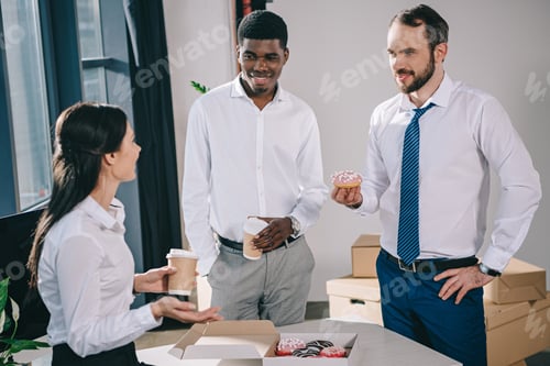 Preview: smiling multiethnic coworkers drinking coffee to go and eating donuts in new office