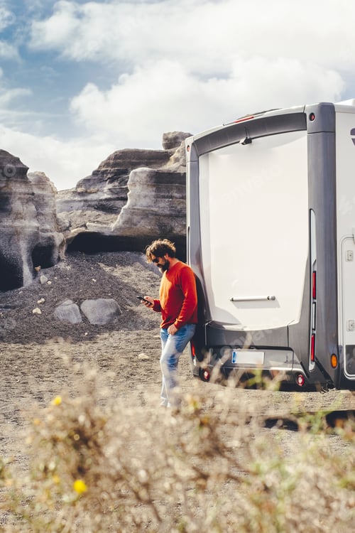 Preview: Standing man using mobile phone against a modern camper van and country side rural landscape in back