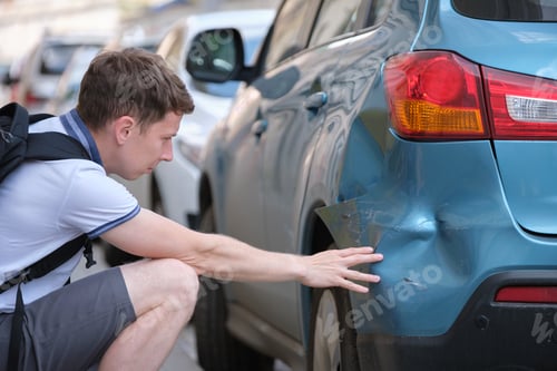 Preview: Sad young man driver sitting near his dented car looking shocked on damaged fender in road accident