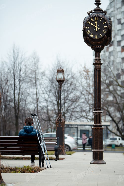 Preview: A young man with a leg injury is sitting on a bench next to a street clock, crutches are nearby.