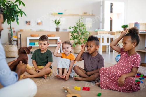 Preview: Nursery school children with man teacher sitting on floor indoors in classroom, raising hands.