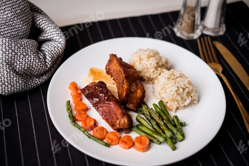 Preview: grilled rib on white plate with rice and asparagus on dark background with gold knife