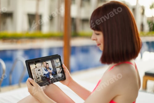 Preview: Woman relaxing at tropical resort pool with tablet pc, having video call with her friends