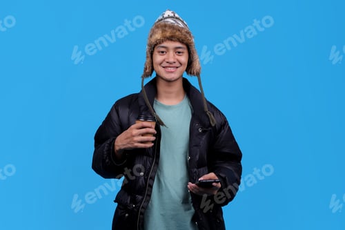 Preview: Smiling young man in a winter hat holding coffee and smartphone on blue background