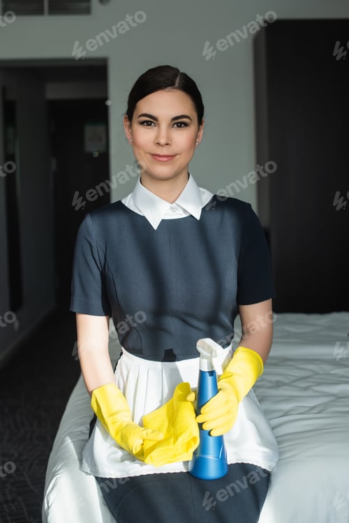 Preview: smiling chambermaid in rubber gloves holding spray bottle and sitting on bed in hotel room