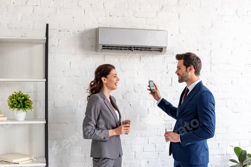 Preview: smiling businesspeople talking while standing under air conditioner in office