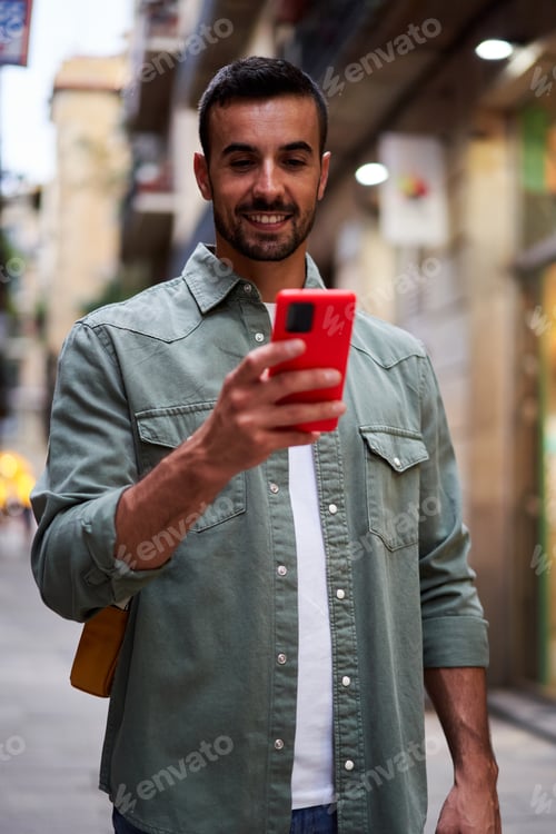 Visualização: Jovem caucasiano alegre segurando o celular parado na rua da cidade.
