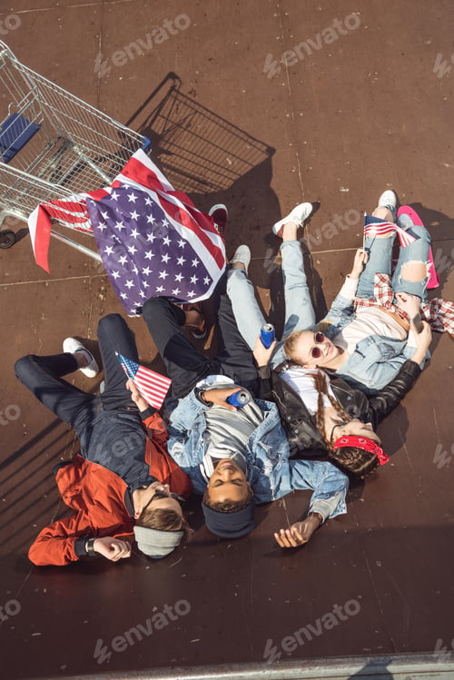 Preview: elevated view of teenagers having fun and lying with american flag in skateboard park, hipster style