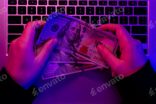Preview: Money in male hands on the background of a laptop keyboard in neon lighting.