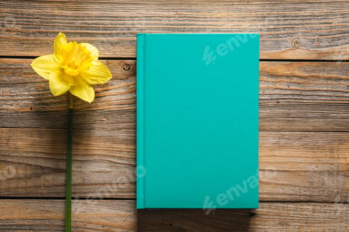 Preview: Closed green book and yellow daffodil flower on wooden background, top view.
