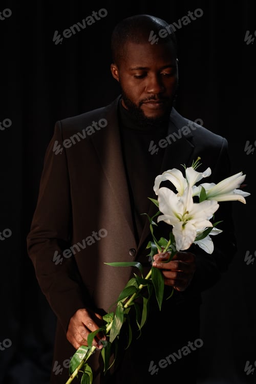 Preview: Black Man in Suit Holding Flower in Studio with Dim Lightning on Black Background