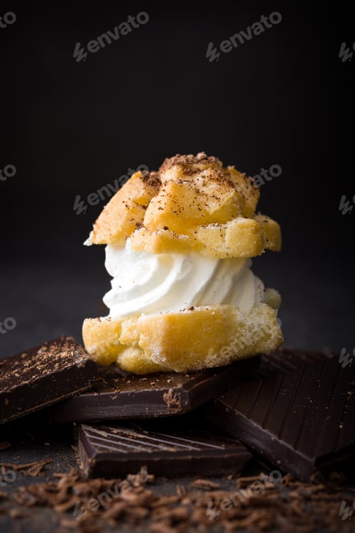 Preview: Cream puff balancing on chocolate chunks with chocolate shavings on dark background