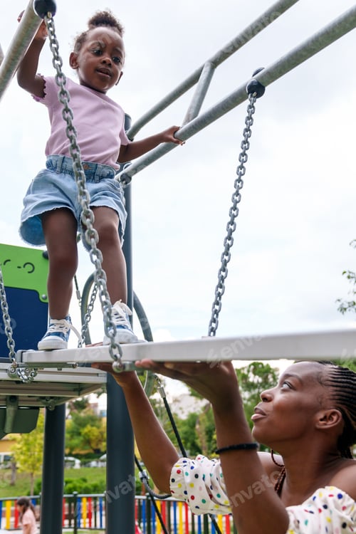 Preview: Fearful African Girl, 5, Crossing Playful Bridge in Colorful Park, Mother Protectively Watching