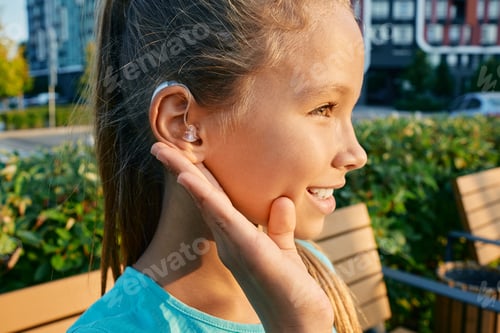 Preview: Smiling female child with a hearing aid behind the ear holds hand near her ear for listening