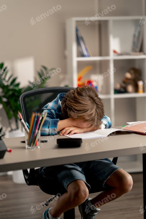 Preview: in the children's room a schoolboy in white sweater and a blue checkered shirt lies on a desk behind