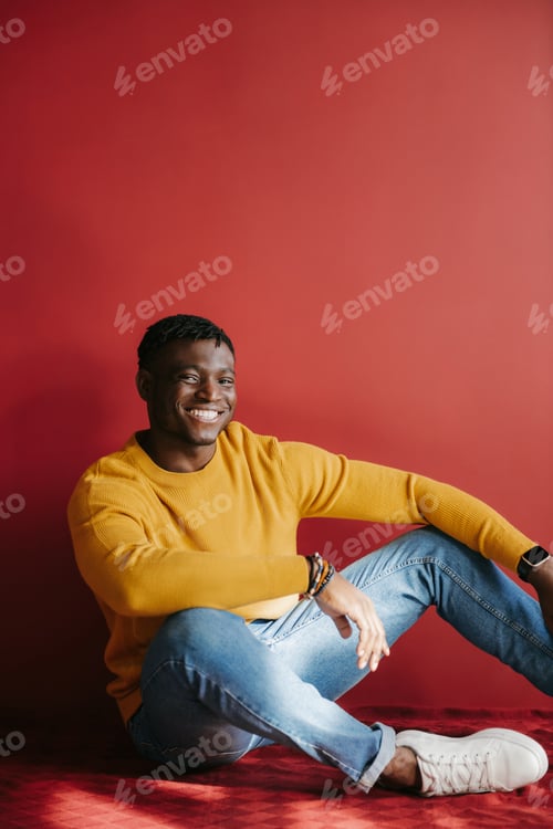Preview: Relaxed young African man in casual wear looking happy while sitting on the floor