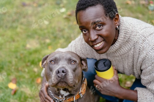 Preview: Black woman with American Staffordshire Terrier sitting on grass holding yellow cup in autumn park