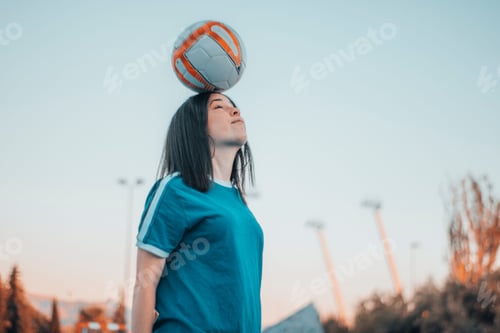 Preview: Closeup of a pretty Caucasian woman in a blue t-shirt holding a soccer ball on her head outdoors