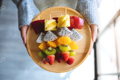 Preview: Top view image of a woman holding a wooden plate of fresh mixed fruits on skewers