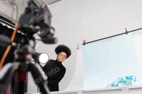 Preview: african american content maker looking at blurred digital camera near spotlight and shooting table