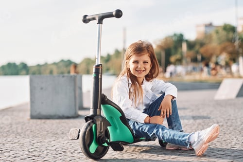 Preview: Sitting and smiling. Cute little girl with scooter is outdoors in summertime
