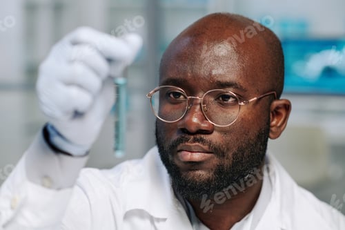 Preview: Young bearded scientist in eyeglasses and gloves looking at flask with liquid