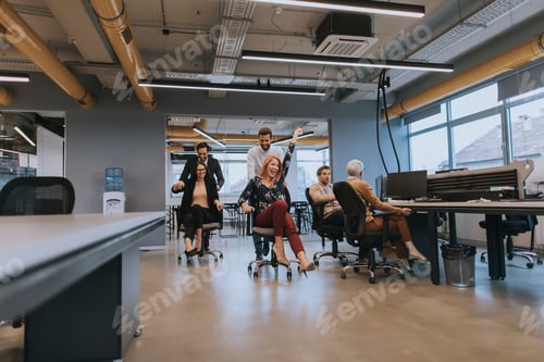 Preview: Young business people in smart casual wear having fun while racing on office chairs and smiling