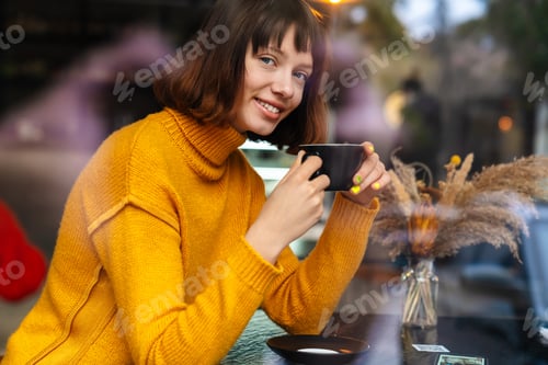 Preview: Happy redhead girl smiling and drinking coffee while sitting in cafe