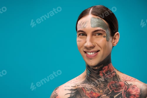 Preview: tattooed young man with clay mask on face smiling isolated on blue