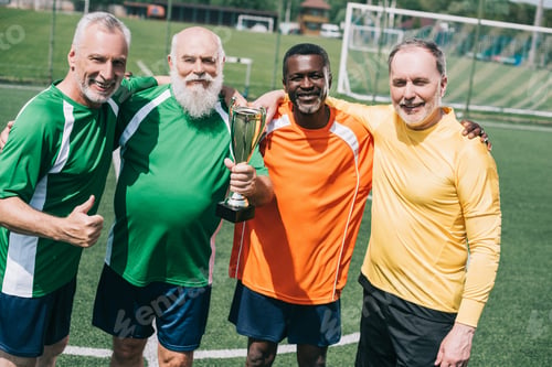 Preview: multiethnic smiling old sportsmen with champions cup standing on football field