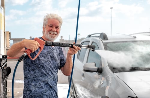 Preview: Smiling senior man washing his car in a self-service car wash station using high pressure pump