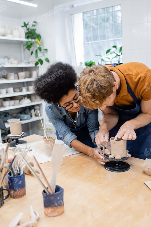 Preview: African american craftswoman helping man making clay cup in pottery studio
