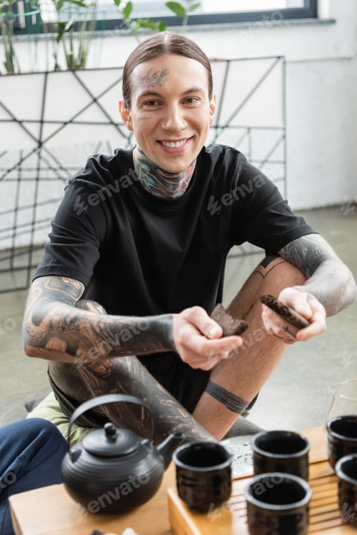 Preview: cheerful young man with tattoos breaking compressed puer tea near traditional Chinese tea pot