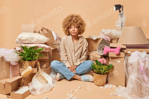 Preview: Thoughtful curly haired young European woman sits crossed legs on floor wears shirt and jeans surrou