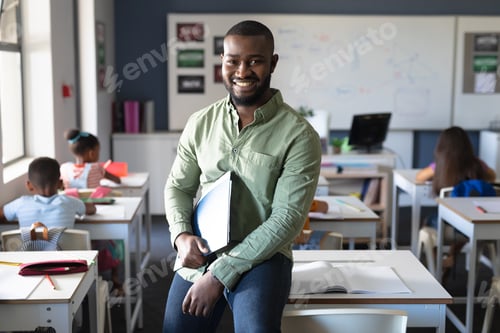 Preview: Portrait of smiling african american young male teacher holding file while sitting at desk in class