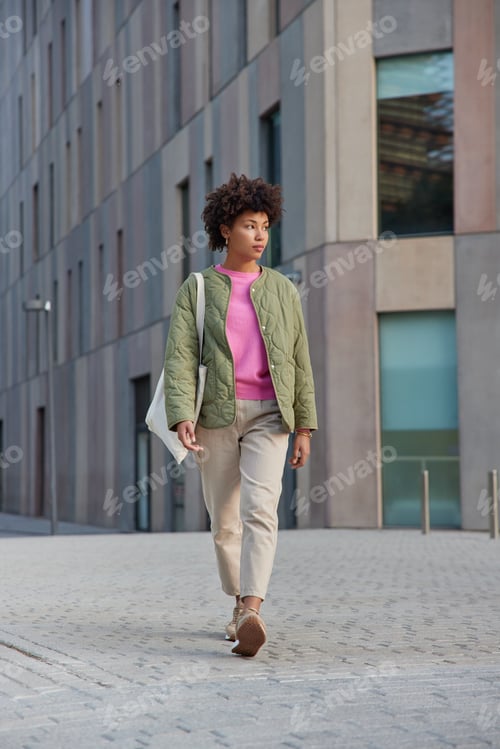 Preview: Full length shot of curly haired woman wears jacket trousers and sneakers carries fabric bag strolls