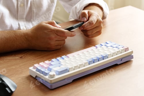 Preview: Person using smartphone while sitting at desk with a colorful keyboard in a bright room