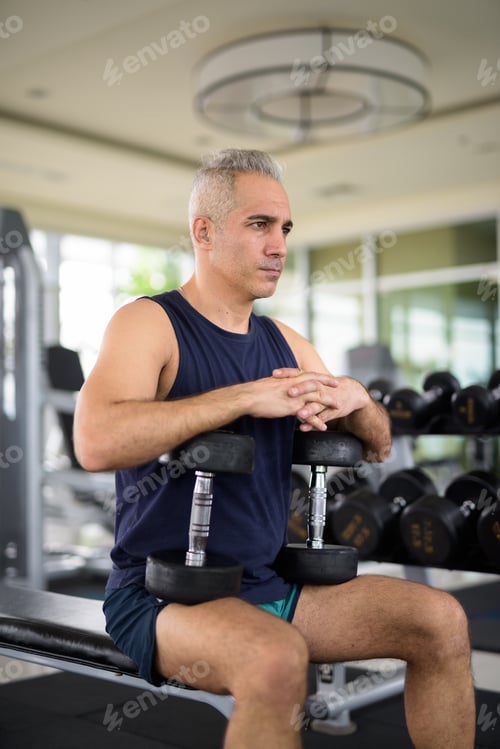 Preview: Mature handsome Persian man exercising with dumbbells and sitting at the gym