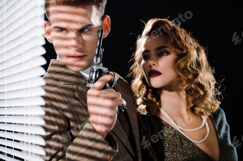 Preview: selective focus of gangster holding gun and looking at window blinds near woman isolated on black