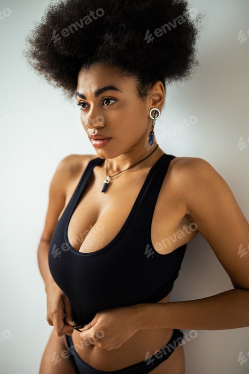 Preview: Curly african american woman in crop top and panties looking away while standing near wall