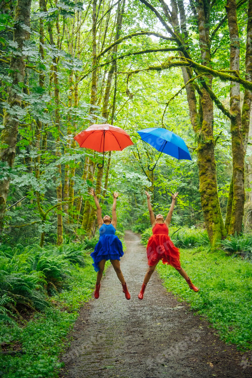 Preview: Two people joyfully leap into the air on a forest path, each holding a colorful umbrella.