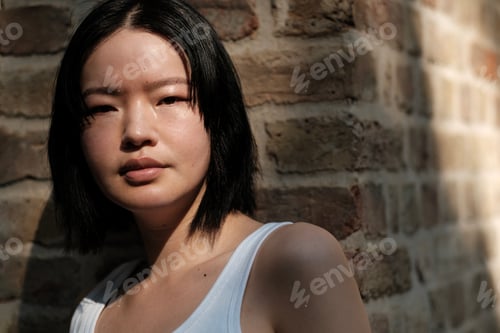 Preview: Young asian woman posing near brick wall in sunlight