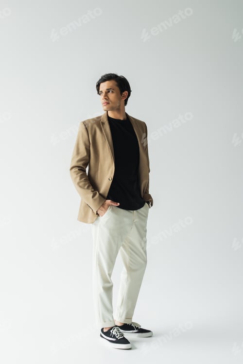Preview: full length of young man in stylish beige blazer standing with hands in pockets on grey