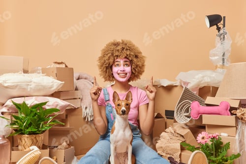 Preview: Positive curly haired woman dressed in casual t shirt and denim overalls points index fingers above